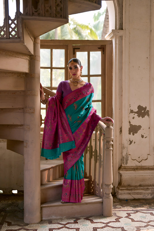 Woman wearing a magenta silk saree with teal pallu and intricate gold floral motifs, paired with a coordinated blouse and traditional gold jewelry, elegantly standing on a vintage spiral staircase in a sunlit heritage interior with tall wooden windows, distressed off-white walls, and patterned tile flooring.
