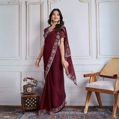 Woman wearing a maroon georgette saree adorned with delicate sequins and an ornate silver embroidered border, paired with a matching blouse and traditional jhumka earrings. She stands elegantly in a classic white paneled room with a vintage rotary telephone on a carved wooden side table, a modern wooden chair with a woven seat, and a patterned rug, creating a sophisticated and timeless ambiance.