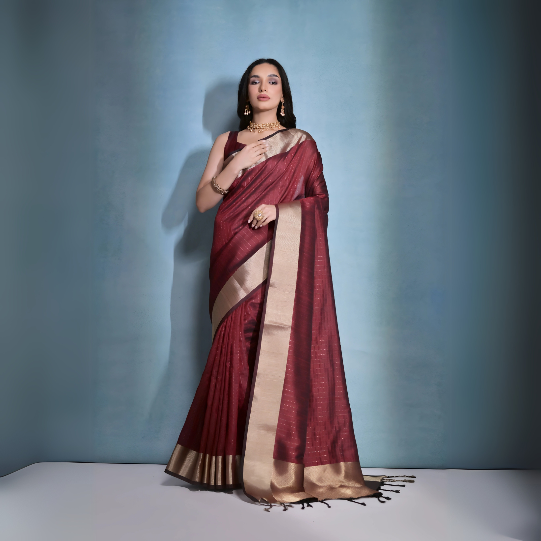 Woman wearing a maroon silk saree with gold border and traditional jewelry, standing against a blue background.