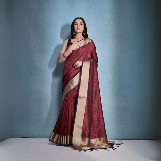 A woman models a striking Dark Maroon Banarasi Raw Silk Saree. The saree features a very wide, luxurious metallic gold Zari border and subtle woven stripes on the body. She wears a sleeveless maroon blouse and traditional gold Kundan jewelry, posing indoors.