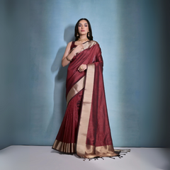 Woman wearing a maroon silk saree with gold border and traditional jewelry, standing against a blue background.