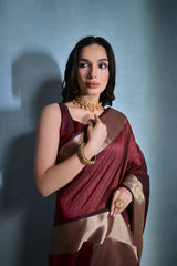 Woman wearing a maroon silk saree with gold border and traditional jewelry, standing against a blue background.