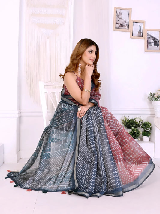 Woman wearing a maroon and grey cotton saree featuring bold geometric and tribal-inspired patterns on the pallu and pleats, paired with a sleeveless blouse and statement silver earrings. She poses elegantly in a contemporary studio setting with white walls, hanging macramé decor, floral arrangements, and framed minimalist artwork, showcasing the saree’s intricate design and graceful drape.