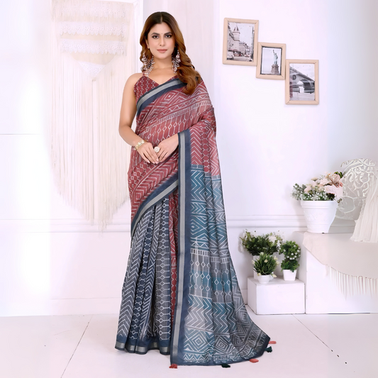 Woman wearing a maroon and grey cotton saree featuring bold geometric and tribal-inspired patterns on the pallu and pleats, paired with a sleeveless blouse and statement silver earrings. She poses elegantly in a contemporary studio setting with white walls, hanging macramé decor, floral arrangements, and framed minimalist artwork, showcasing the saree’s intricate design and graceful drape.