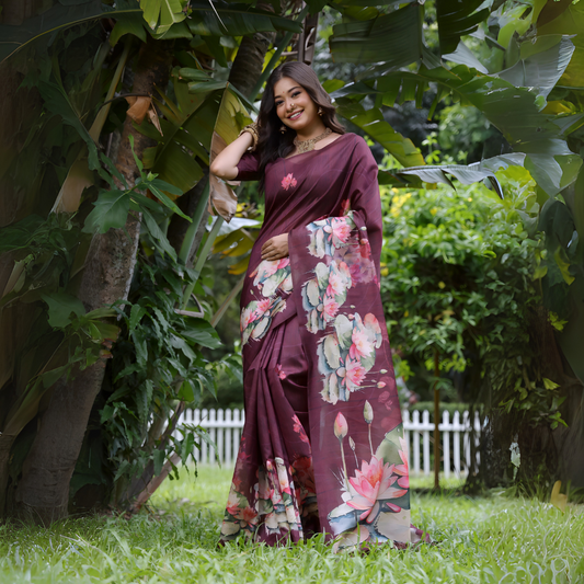 Woman wearing a maroon saree with large handpainted lotus and leaf floral motifs, accessorized with traditional jewelry, posing gracefully outdoors amid lush tropical greenery and a white picket fence in the background