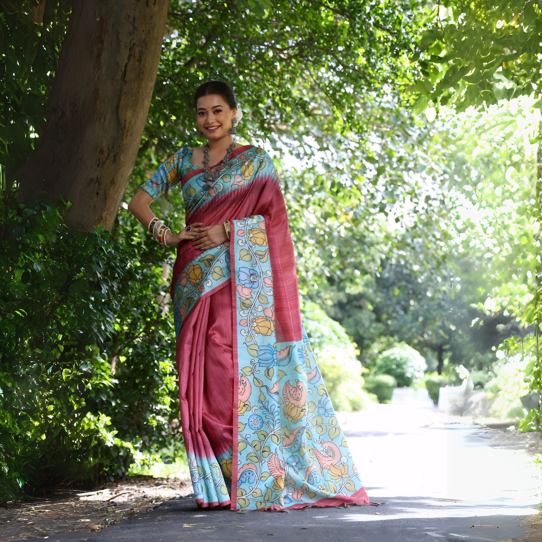 Woman wearing a maroon saree with vibrant Madhubani-inspired floral print border and pallu, paired with a matching blouse and traditional jewelry, standing gracefully on a sunlit garden pathway surrounded by lush green trees and foliage.
