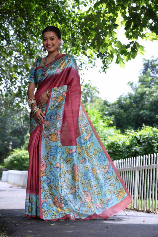 Woman wearing a maroon saree with vibrant Madhubani-inspired floral print border and pallu, paired with a matching blouse and traditional jewelry, standing gracefully on a sunlit garden pathway surrounded by lush green trees and foliage.
