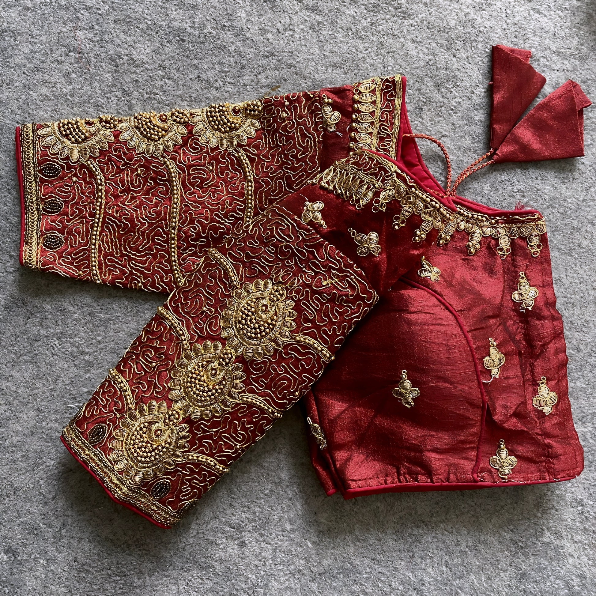 Traditional maroon silk blouse featuring intricate gold embroidery and beadwork with floral and paisley motifs on the sleeves and neckline, displayed flat on a textured surface with a bunch of artificial flowers and a woven mat in the background.