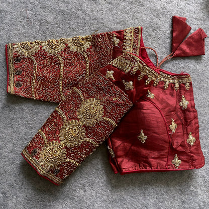 Traditional maroon silk blouse featuring intricate gold embroidery and beadwork with floral and paisley motifs on the sleeves and neckline, displayed flat on a textured surface with a bunch of artificial flowers and a woven mat in the background.