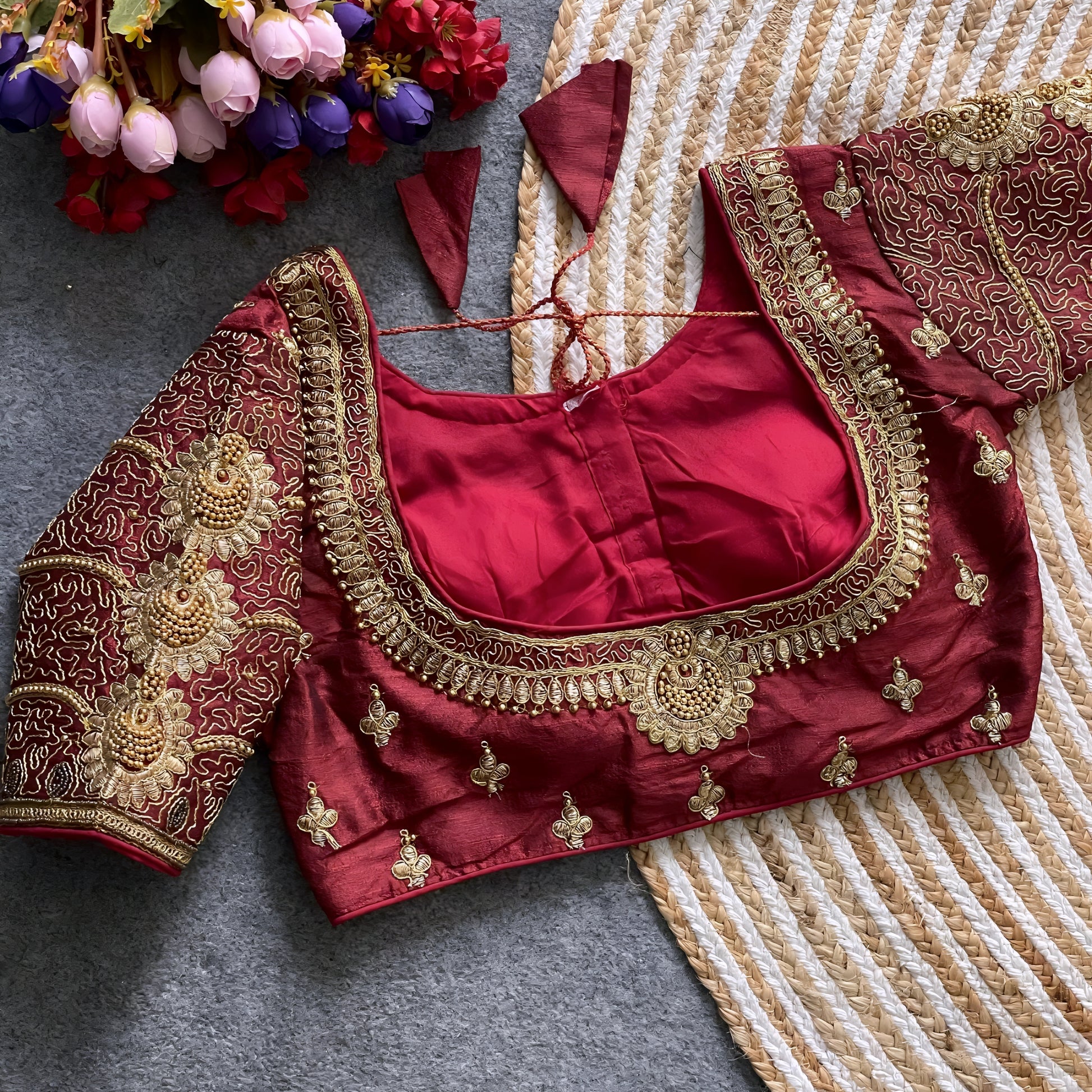 Traditional maroon silk blouse featuring intricate gold embroidery and beadwork with floral and paisley motifs on the sleeves and neckline, displayed flat on a textured surface with a bunch of artificial flowers and a woven mat in the background.