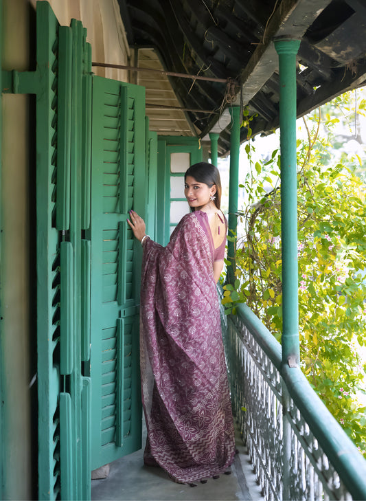 Woman wearing a mauve floral printed saree with a matching blouse, standing on a vintage balcony with green shutters and railings, surrounded by leafy vines and natural light.