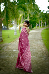 Woman wearing a mauve handloom cotton saree with white tribal-inspired prints and matching blouse, accessorized with traditional jewelry, standing on a stone pathway surrounded by lush green palm trees in a garden setting.