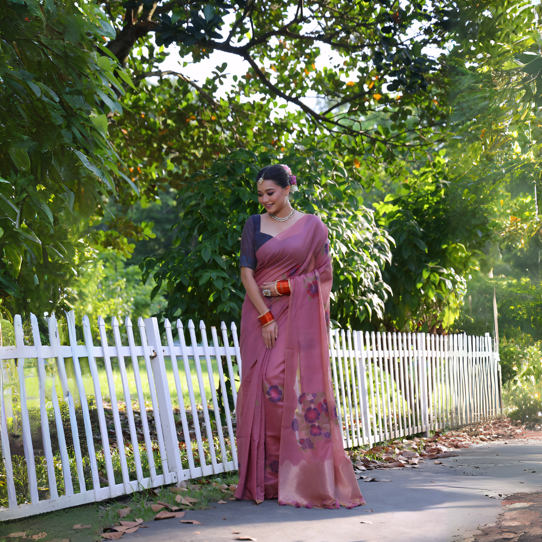 Woman wearing a mauve silk saree with subtle floral embroidery and a navy blue blouse, accessorized with traditional jewelry and red bangles, standing beside a white picket fence in a sunlit garden surrounded by lush green trees.