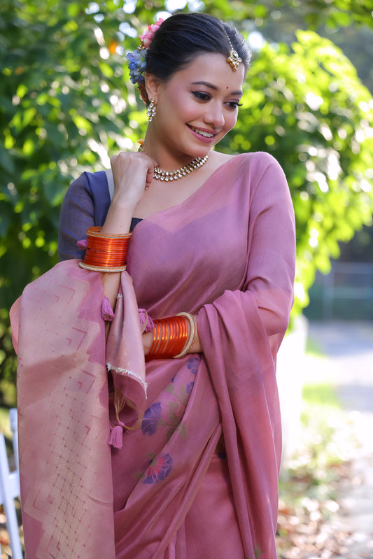 A woman models an elegant Dusty Rose Pink Soft Muga Cotton Saree. The pallu features large, colorful Resham floral weaving in purple and dark pink. She wears a slate grey blouse and is accessorized with a pearl choker necklace and vibrant orange bangles, posing outdoors by a white fence.