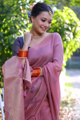 Woman wearing a mauve silk saree with subtle floral embroidery and a navy blue blouse, accessorized with traditional jewelry and red bangles, standing beside a white picket fence in a sunlit garden surrounded by lush green trees.