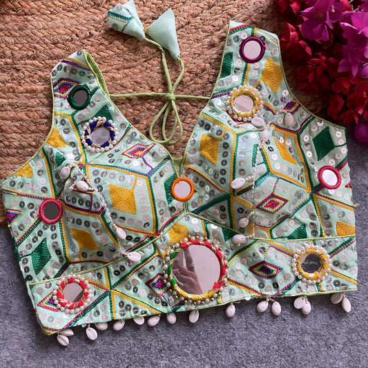 Traditional mint green Gujarati-style blouse featuring vibrant multicolor geometric embroidery with large circular mirrors bordered by colorful beads, decorative cowrie shell trim along the hem, and tasseled tie-back detail. Displayed flat on a grey carpet with a woven mat and bold fuchsia artificial flowers in the corner.