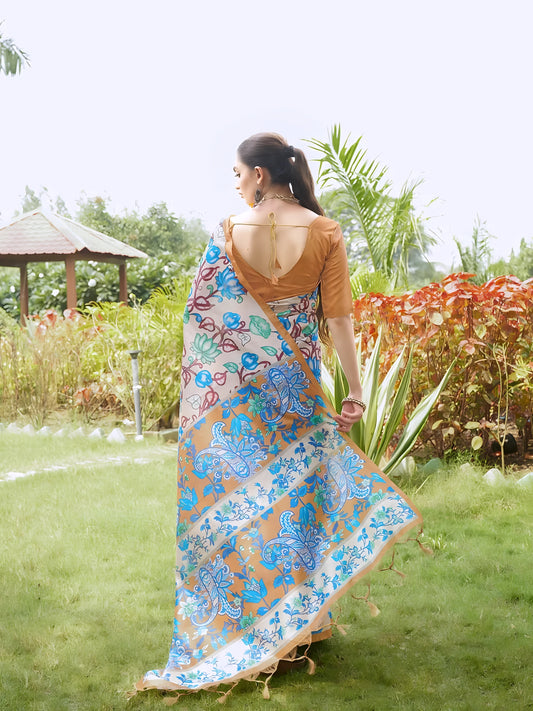 Woman wearing a multicolor Madhubani-printed saree with vibrant floral and abstract motifs, paired with a brown blouse and traditional jewelry, standing gracefully on a lush green lawn surrounded by tropical plants and red foliage, with a garden gazebo in the background.