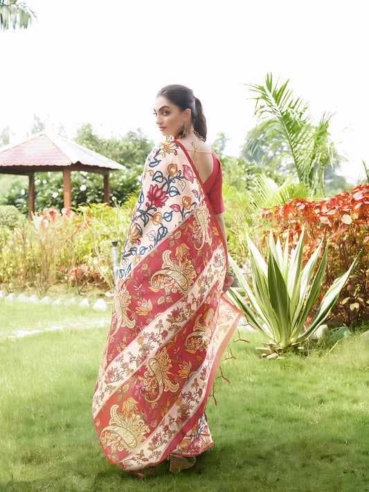 Woman wearing a multicolor Madhubani-printed saree with a vibrant red blouse and traditional jewelry, standing gracefully on a lush green lawn with landscaped garden plants and a red-roofed gazebo in the background.