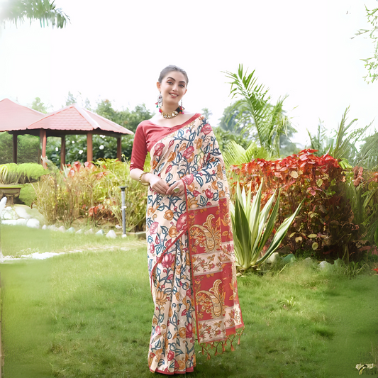 Woman wearing a multicolor Madhubani-printed saree with a vibrant red blouse and traditional jewelry, standing gracefully on a lush green lawn with landscaped garden plants and a red-roofed gazebo in the background.
