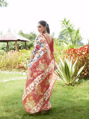 Woman wearing a multicolor Madhubani-printed saree with a vibrant red blouse and traditional jewelry, standing gracefully on a lush green lawn with landscaped garden plants and a red-roofed gazebo in the background.