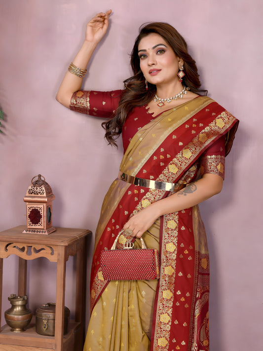 Woman draped in a mustard yellow and deep red Banarasi silk saree featuring intricate gold woven motifs and an ornate red and gold embroidered border, paired with a matching red blouse with gold detailing. She poses gracefully in a contemporary studio with blush-toned walls, rustic white vases, a wooden side table adorned with brass decor, green foliage, and a bowl of yellow flowers on the floor.