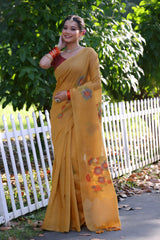 Woman wearing a mustard yellow silk saree with subtle floral embroidery and a contrasting maroon blouse, accessorized with traditional jewelry and bangles, standing beside a white picket fence in a sunlit garden with lush greenery and trees in the background.
