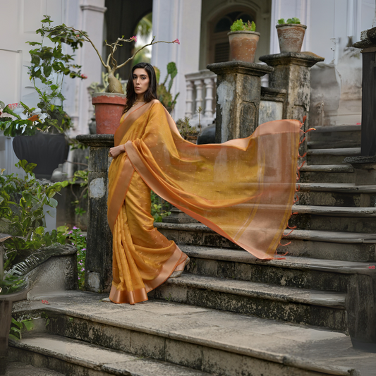 Woman wearing a mustard yellow silk saree with a gold border and delicate woven patterns, gracefully posing on the stone steps of a heritage mansion surrounded by potted plants and lush greenery.