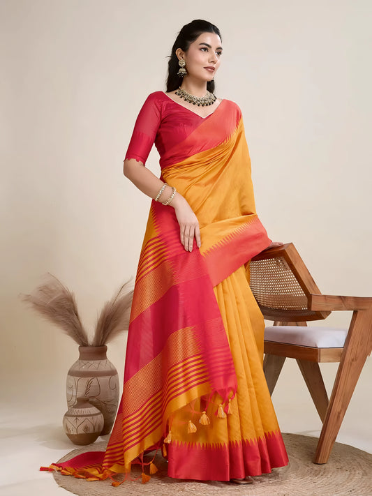 Woman wearing a mustard yellow silk saree with a bold red border and pallu, paired with a matching red blouse and traditional jewelry, elegantly posing beside a wooden chair in a minimalist studio setting with neutral decor, ceramic vases, and pampas grass.Woman wearing a mustard yellow silk saree with a bold red border and pallu, paired with a matching red blouse and traditional jewelry, elegantly posing beside a wooden chair in a minimalist studio setting with neutral decor, ceramic vases, and pampas gras