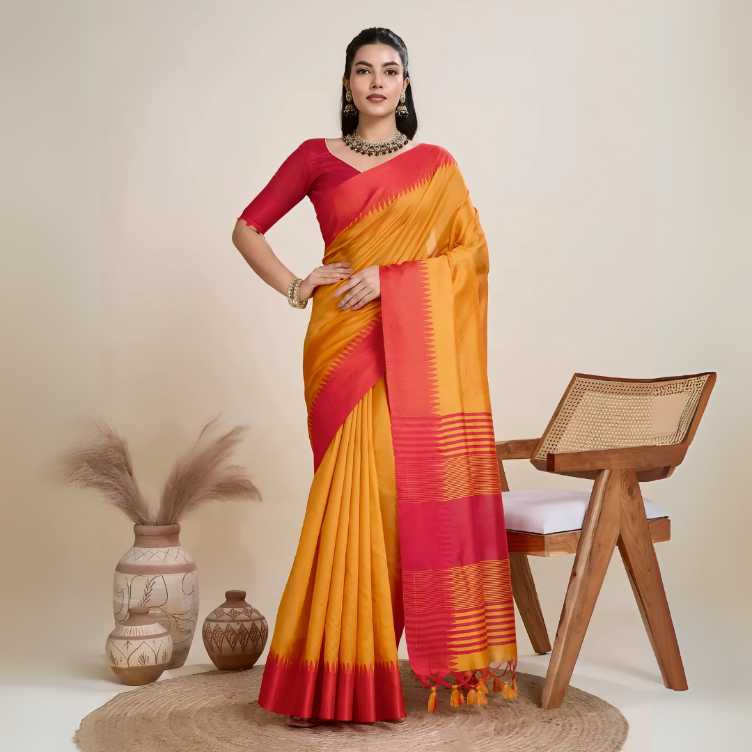 Woman wearing a mustard yellow silk saree with a bold red border and pallu, paired with a matching red blouse and traditional jewelry, elegantly posing beside a wooden chair in a minimalist studio setting with neutral decor, ceramic vases, and pampas grass.