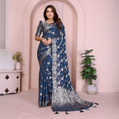 Woman wearing a navy blue Banarasi saree adorned with silver zari motifs and a rich traditional border, paired with a matching silver blouse with short sleeves. Styled with elegant jewelry and bangles, she stands in front of a pastel arched backdrop with minimal decor, including a white side table with vases and a potted green plant, presenting a graceful festive ethnic look.