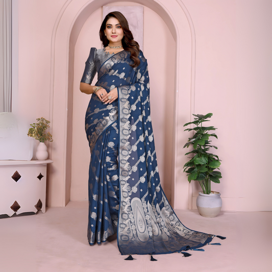 Woman wearing a navy blue Banarasi saree adorned with silver zari motifs and a rich traditional border, paired with a matching silver blouse with short sleeves. Styled with elegant jewelry and bangles, she stands in front of a pastel arched backdrop with minimal decor, including a white side table with vases and a potted green plant, presenting a graceful festive ethnic look.