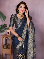 Woman wearing a navy blue Banarasi silk saree featuring an ornate golden zari border with traditional circular woven motifs, paired with a matching metallic blouse and gold jewelry. She poses elegantly in a modern studio decorated with large rustic white clay pots, a wooden stand displaying brass decorative items, a bowl of fresh marigold flowers, and green potted foliage, all set against a soft mauve backdrop.
