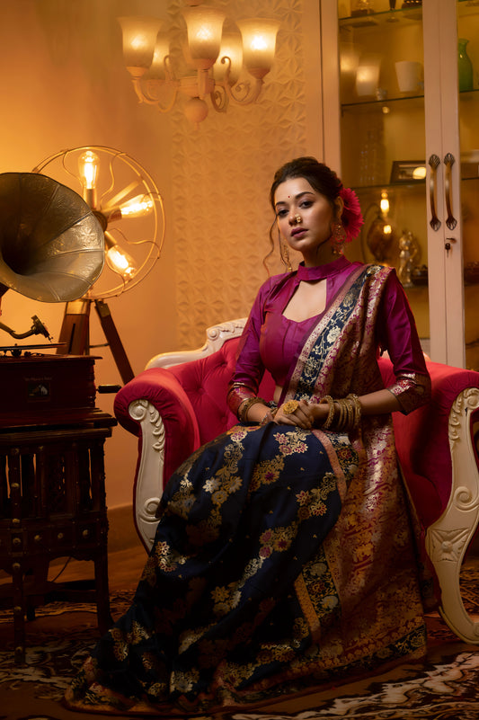 Woman wearing a navy blue Banarasi silk saree with intricate gold and pink zari work, paired with a magenta blouse and traditional jewelry, standing beside a vintage gramophone in an elegant, warmly lit room with a chandelier and glass display cabinet.