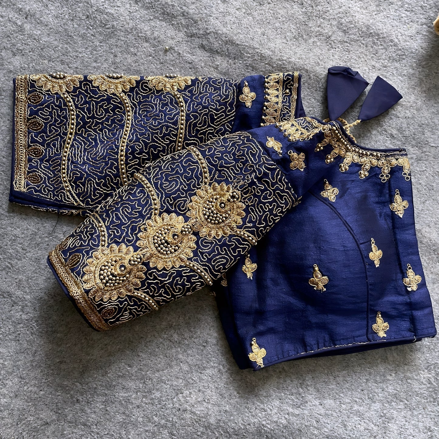Traditional navy blue silk blouse featuring intricate gold embroidery and beadwork with floral and paisley motifs on the sleeves and neckline, displayed flat on a textured surface with a bunch of artificial flowers and a woven mat in the background.