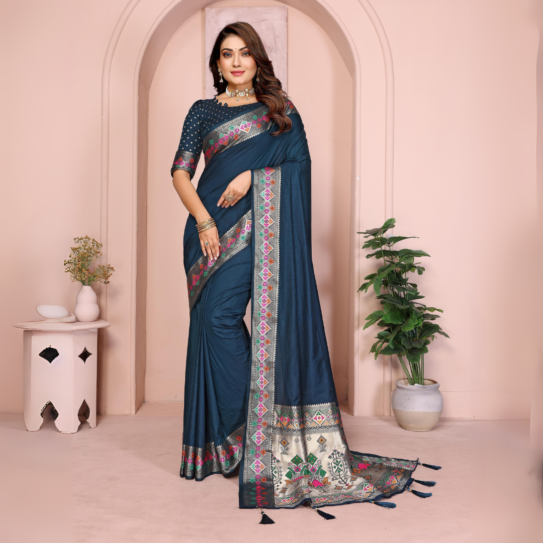  Woman wearing a navy blue silk saree featuring a richly embroidered border with floral and geometric patterns in pink, green, and gold, paired with a matching blouse with silver polka dot embroidery. She accessorizes with a traditional choker necklace, bangles, and rings, posed elegantly in a pastel arched alcove studio with minimalist decor, a white side table, and potted green plants.