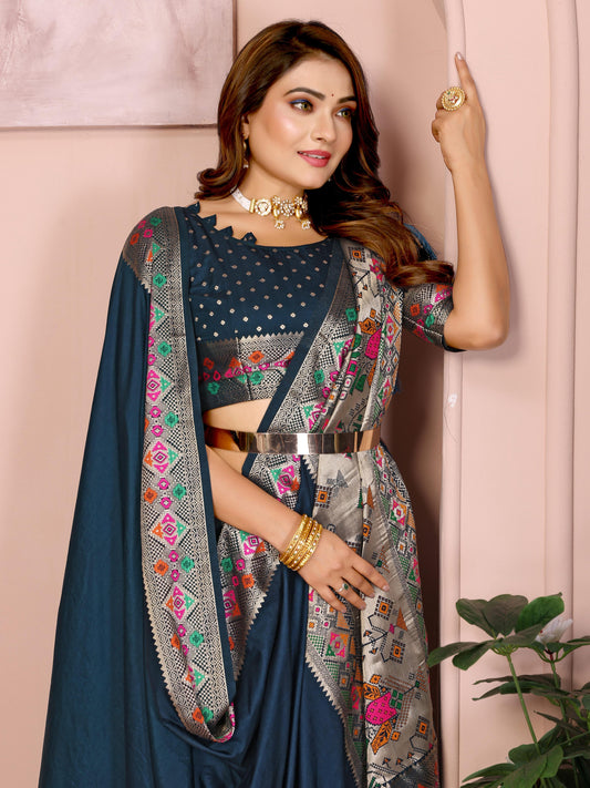  Woman wearing a navy blue silk saree featuring a richly embroidered border with floral and geometric patterns in pink, green, and gold, paired with a matching blouse with silver polka dot embroidery. She accessorizes with a traditional choker necklace, bangles, and rings, posed elegantly in a pastel arched alcove studio with minimalist decor, a white side table, and potted green plants.