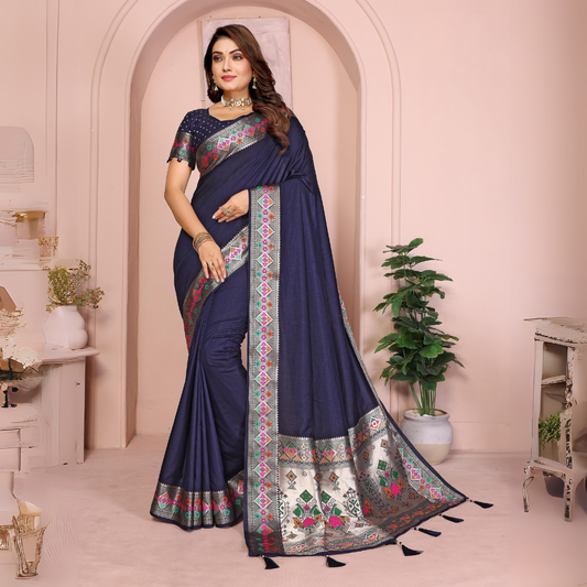 Woman wearing a navy blue silk saree featuring a richly embroidered border with floral and geometric patterns in pink, green, and gold, paired with a matching blouse with silver polka dot embroidery. She accessorizes with a traditional choker necklace, bangles, and rings, posed elegantly in a pastel arched alcove studio with minimalist decor, a white side table, and potted green plants.