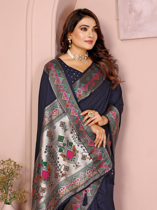 Woman wearing a navy blue silk saree featuring a richly embroidered border with floral and geometric patterns in pink, green, and gold, paired with a matching blouse with silver polka dot embroidery. She accessorizes with a traditional choker necklace, bangles, and rings, posed elegantly in a pastel arched alcove studio with minimalist decor, a white side table, and potted