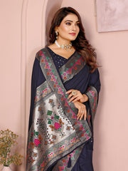 Woman wearing a navy blue silk saree featuring a richly embroidered border with floral and geometric patterns in pink, green, and gold, paired with a matching blouse with silver polka dot embroidery. She accessorizes with a traditional choker necklace, bangles, and rings, posed elegantly in a pastel arched alcove studio with minimalist decor, a white side table, and potted