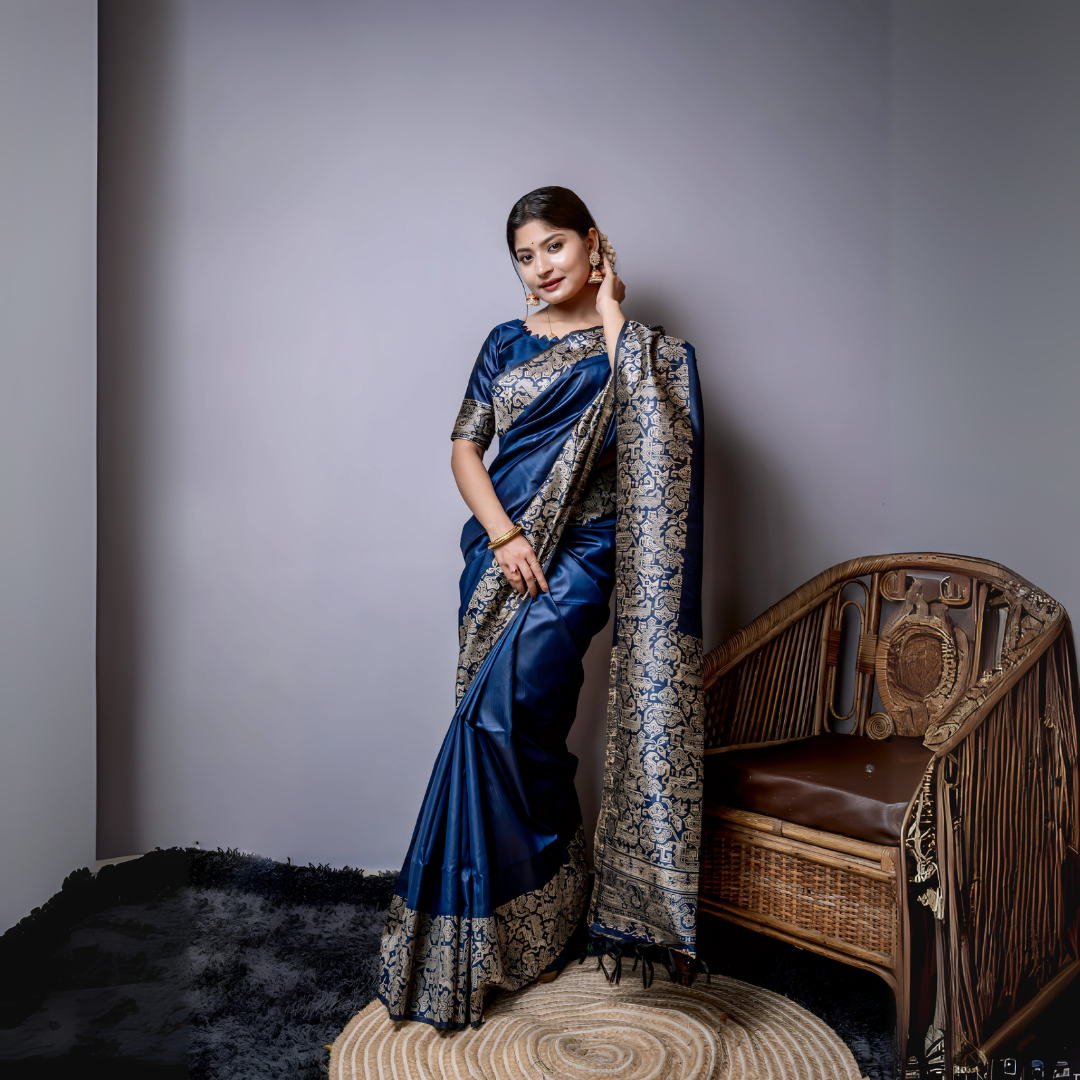 Woman wearing a navy blue silk saree with intricate silver brocade border and pallu, paired with a matching blouse and gold bangles, standing gracefully beside a vintage wooden chair in a minimalist modern studio with grey walls and a textured rug.