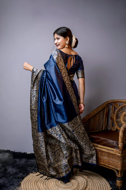 Woman wearing a navy blue silk saree with intricate silver brocade border and pallu, paired with a matching blouse and gold bangles, standing gracefully beside a vintage wooden chair in a minimalist modern studio with grey walls and a textured rug.