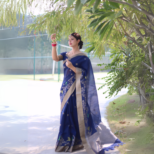 Woman wearing a navy blue saree with gold border and floral embroidery, posing outdoors under leafy trees on a sunlit pathway.