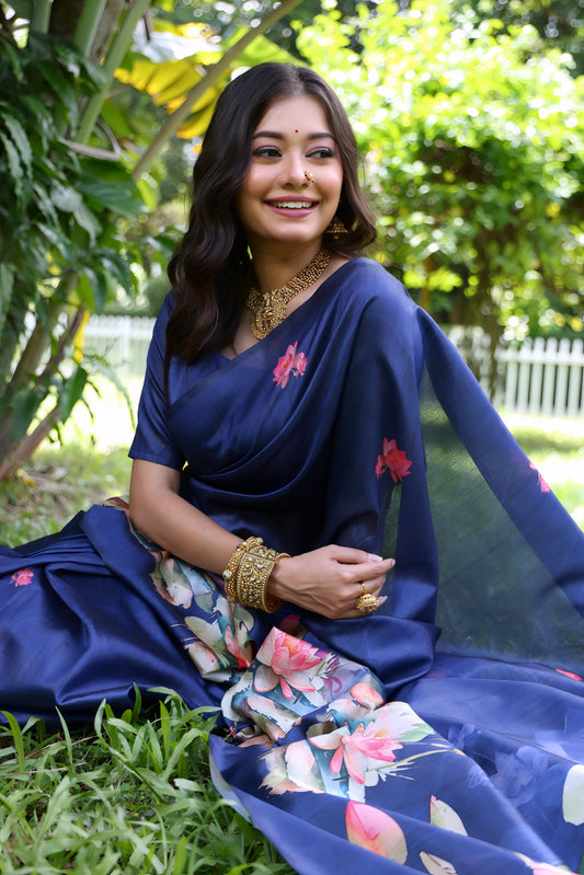 Woman wearing a navy blue saree with large handpainted lotus and leaf floral motifs, accessorized with traditional gold jewelry and bangles, standing on a lush green lawn beneath tropical trees with a white picket fence in the background.