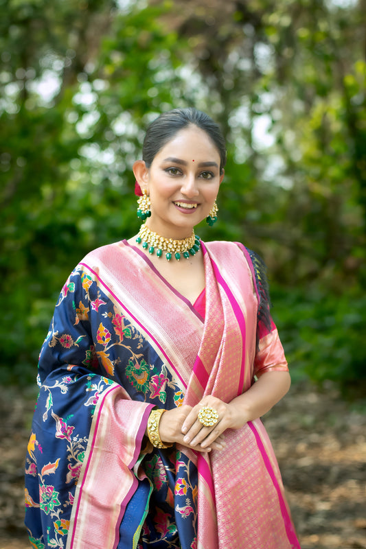  Woman wearing a navy blue silk saree with vibrant multicolor floral motifs and a contrasting pink border, paired with a pink blouse and traditional jewelry, standing gracefully on a shaded garden path surrounded by lush greenery and trees.