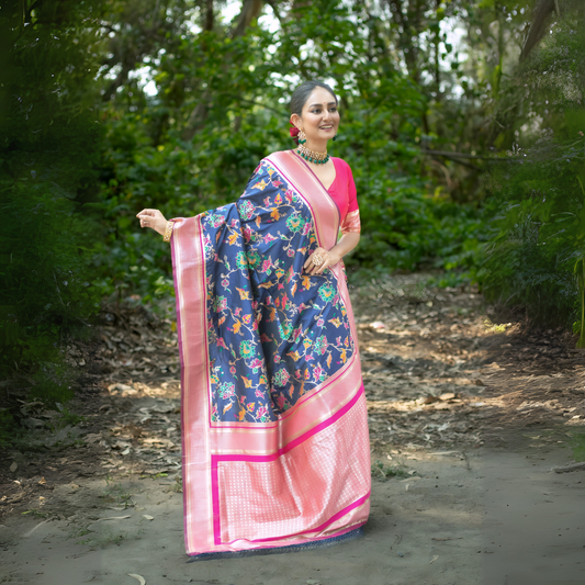  Woman wearing a navy blue silk saree with vibrant multicolor floral motifs and a contrasting pink border, paired with a pink blouse and traditional jewelry, standing gracefully on a shaded garden path surrounded by lush greenery and trees.