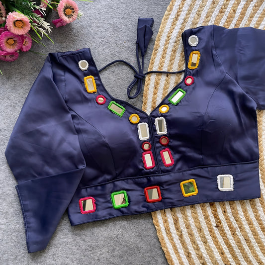 Navy blouse decorated with square and round mirror work featuring multicolored embroidered borders, stylish back tie-up closure, and three-quarter sleeves. Displayed flat on a textured grey surface with a woven mat and pink flowers, highlighting its festive and contemporary ethnic style.