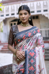 Woman wearing an off-white silk saree with blue and red Patola-inspired geometric and floral borders, featuring intricate floral motifs, paired with a matching blouse and traditional jewelry, elegantly posing on stone steps in front of a vintage heritage house with ornate wooden railings and classic architecture.