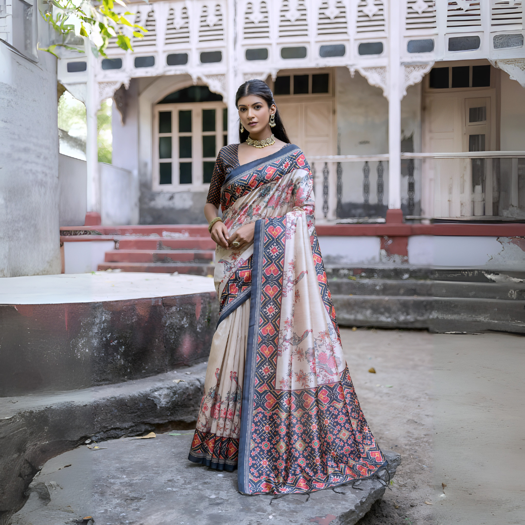 Woman wearing an off-white silk saree with blue and red Patola-inspired geometric and floral borders, featuring intricate floral motifs, paired with a matching blouse and traditional jewelry, elegantly posing on stone steps in front of a vintage heritage house with ornate wooden railings and classic architecture.Woman wearing an off-white silk saree with blue and red Patola-inspired geometric and floral borders, featuring intricate floral motifs, paired with a matching blouse and traditional jewelry, elegan