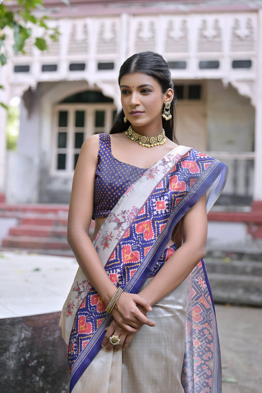 Woman wearing an off-white silk saree with blue and red Patola-inspired geometric and floral patterns, paired with a matching blue blouse and traditional jewelry, elegantly posing in a vintage courtyard with stone steps, ornate pillars, and a heritage building in the background.