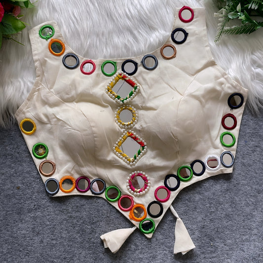 Off-white ethnic blouse featuring vibrant mirror work with multicolored embroidered borders and decorative square and round mirrors arranged vertically at the center, accented with pearl detailing and tie-up tassels at the bottom. Displayed flat on a grey surface with a white faux fur background and green leafy accents, showcasing its festive and traditional design.